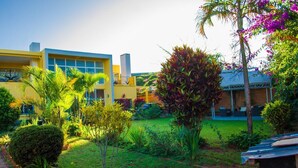 A modern building with a yellow facade and large windows, surrounded by well-maintained greenery and a small palm tree. - Room-the White-orange Bedroom With a ple (Antananarivo)