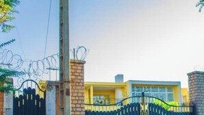 A gated entrance with a black gate featuring yellow lettering, a brick wall, and a yellow building in the background. - Room-the White-orange Bedroom With a ple (Antananarivo)