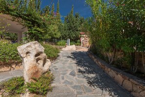 Un chemin de pierres serpente à travers un jardin où l’on trouve une grande sculpture rocheuse et un mur de pierres sur un côté.