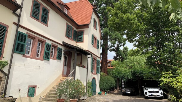 A two-story building with a red-tiled roof, green shutters, and a staircase leading to the entrance.