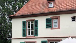 A two-story house with a red-tiled roof, green shutters, and a patio with a table and chairs.