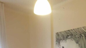 Una habitación de hotel con una cama, un edredón rojo, una almohada, una lámpara fijada a la pared y dos cuadros enmarcados.