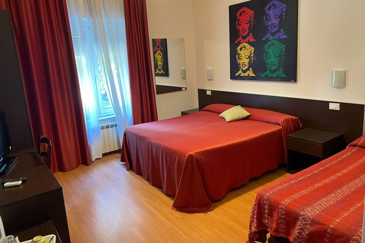 A hotel room with a large bed, a desk, and a chair. There is a window with curtains, a painting on the wall, and a light fixture above the bed.