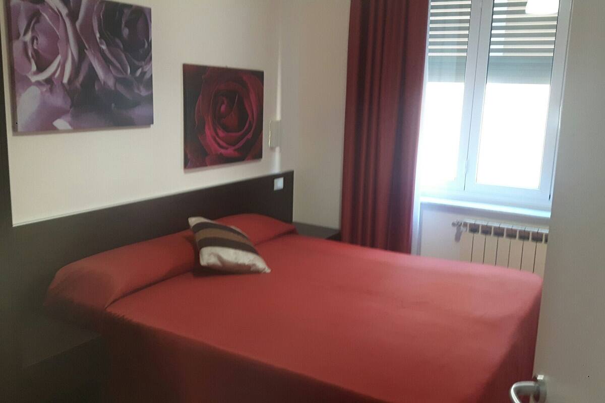 A single bed with a red bedspread and a striped pillow, two framed artworks on the wall, and a window with red curtains.