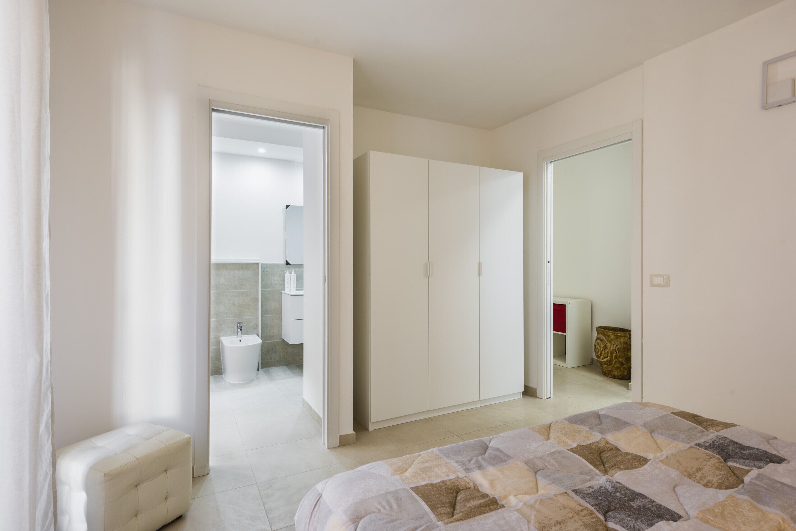 Ein Schlafzimmer mit Bett, Kleiderschrank und einem durchgegangenen Tür erkennbaren Badezimmer sowie einem kleinen Hocker.