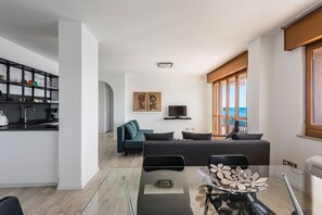 A modern living room with a sofa, a dining table with chairs, a kitchen area with a sink and shelves, and a television.