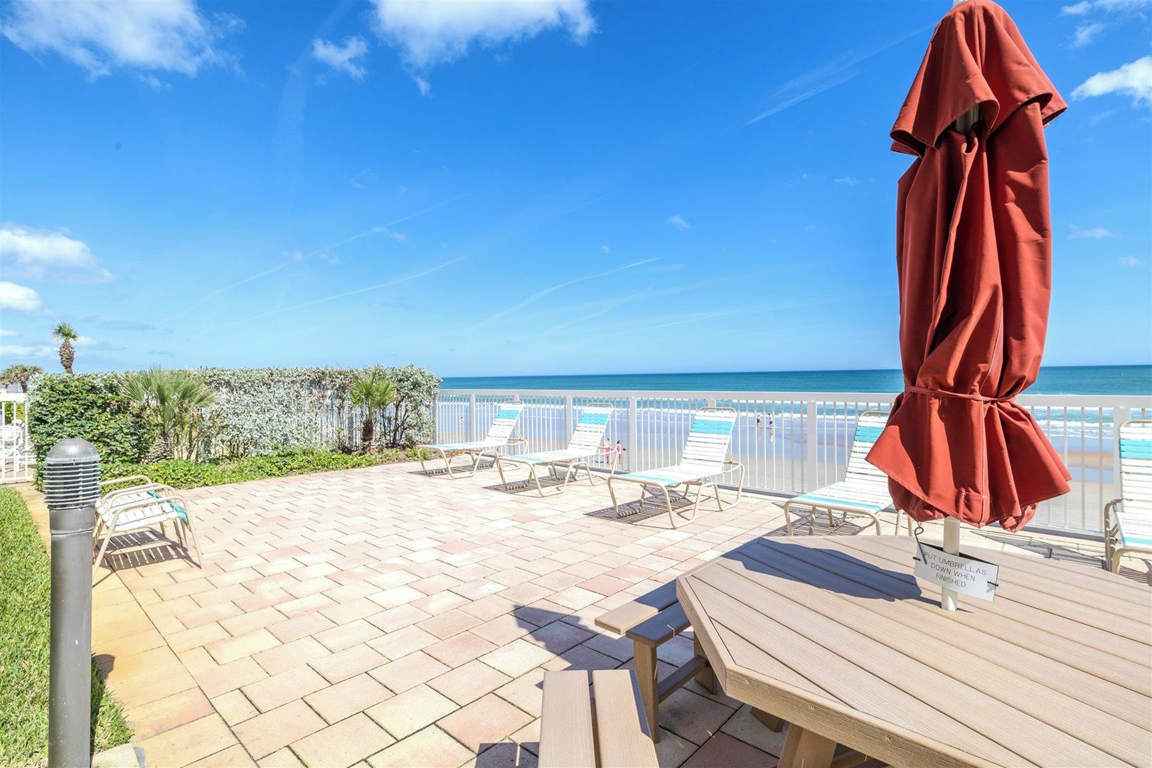 A patio with lounge chairs and a wooden table, overlooking a beach with a clear sky.