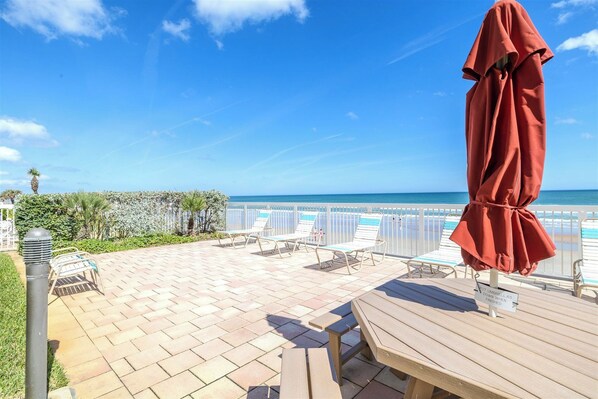 A patio with lounge chairs and a wooden table, overlooking a beach with a clear sky.