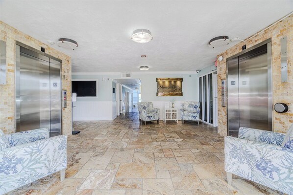 A hotel lobby with two elevators, a long hallway, and a seating area with patterned sofas.