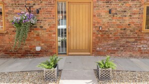 A brick building with a wooden door, a hanging flower basket, and two potted plants on the ground. - The Grange Cottage 1 (Grimsby)