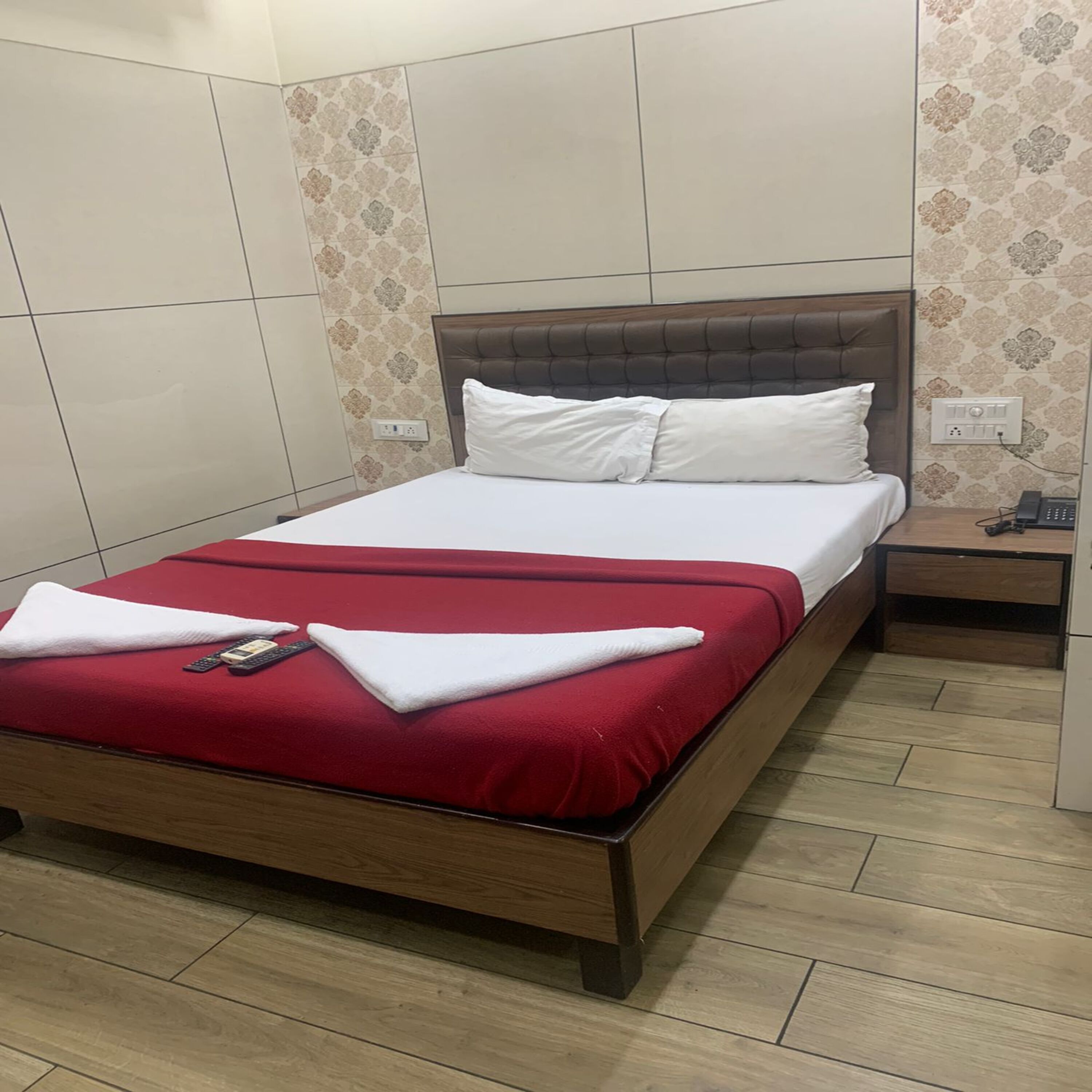 A hotel room with a wooden bed, white bedding, a red throw blanket, a bedside table with a phone, and patterned wallpaper.