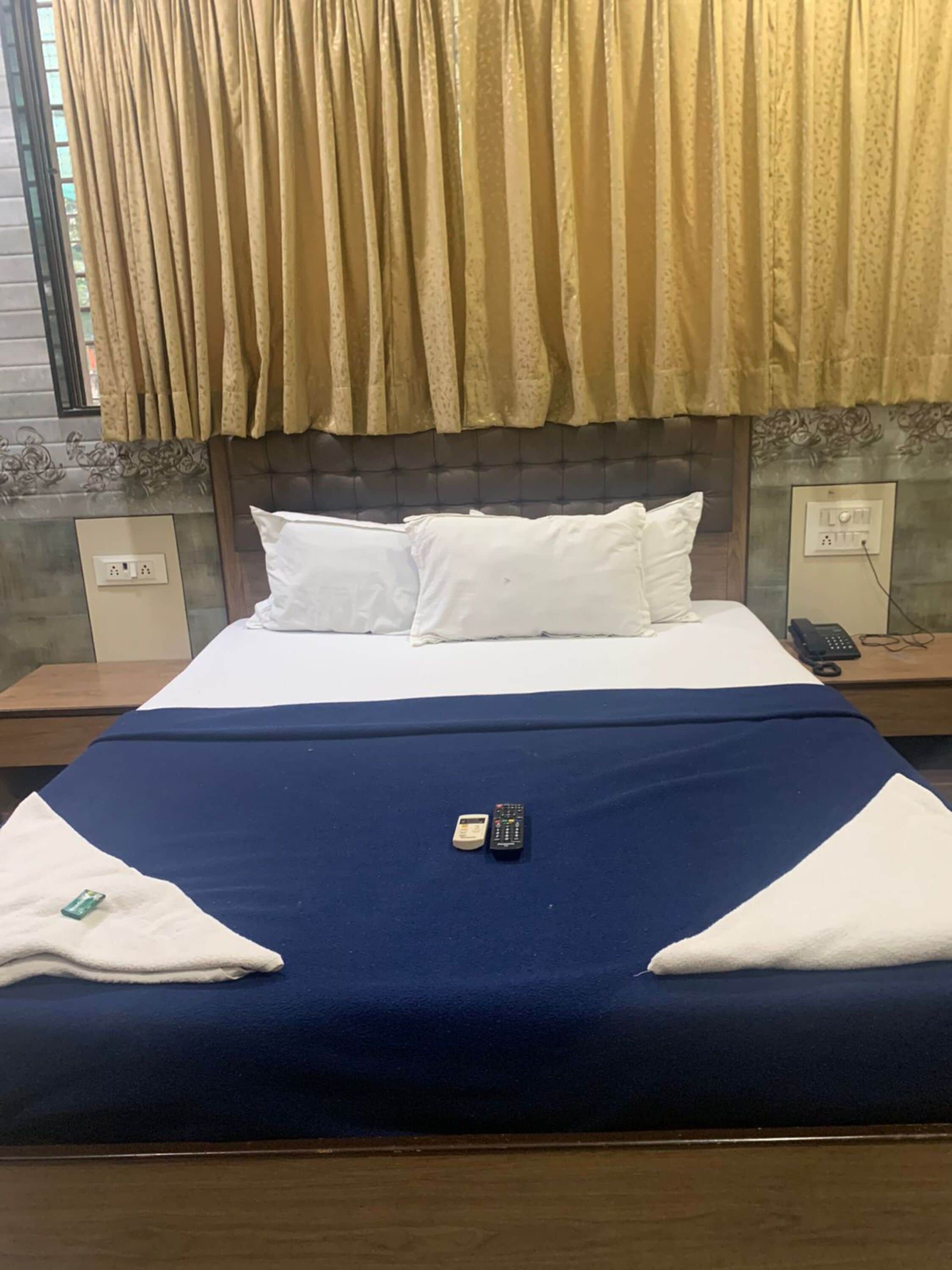 A neatly made bed with a blue bedspread, white pillows, and two remote controls on the bed.