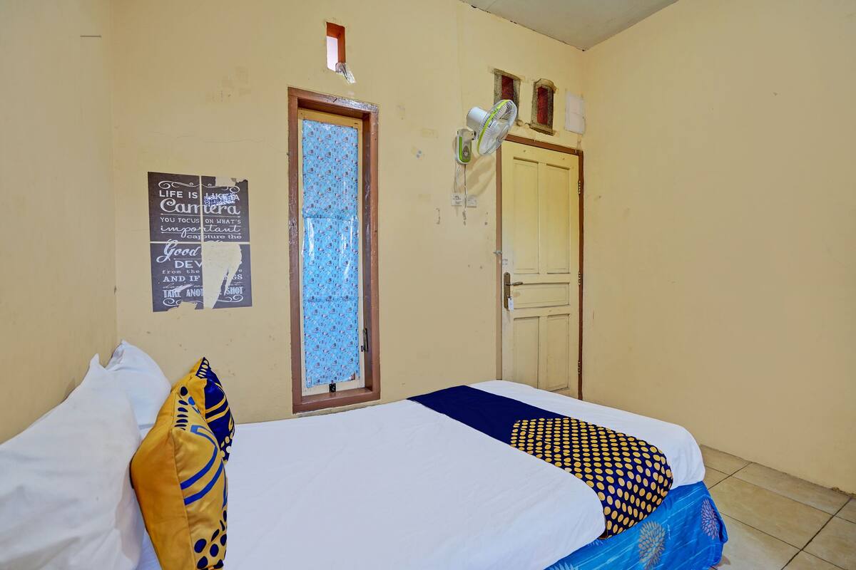 Een kamer met een bed, een raam met een gordijn met patroon, een ventilator aan de muur en een deur.