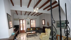 A living room with a sectional sofa, a coffee table, and a patterned armchair. There is a television on a stand, a window with curtains, and a ceiling with exposed beams. - Angelina's House (Rethymno)