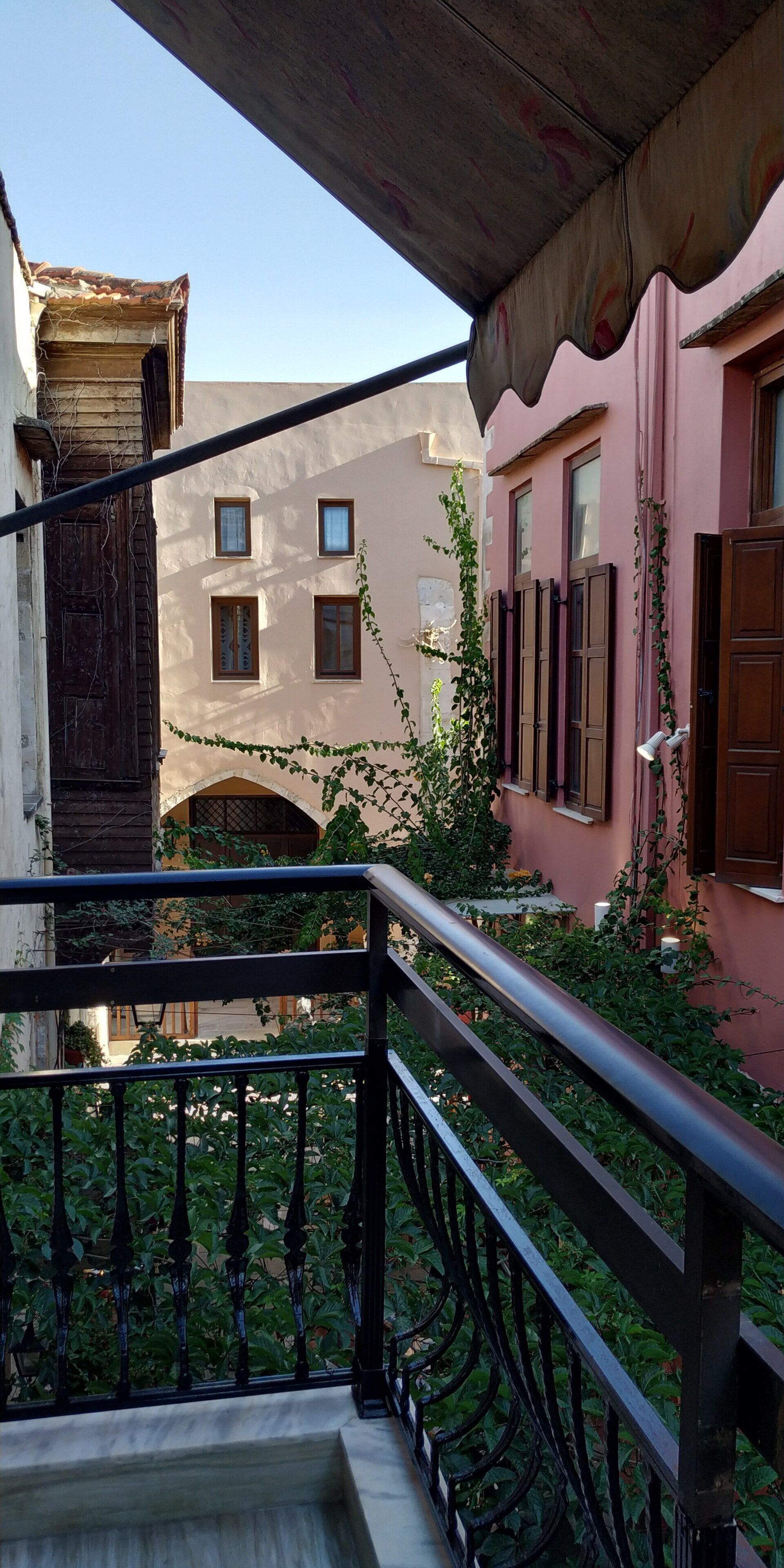Ein Balkon mit schmiedeeisernem Geländer, der auf einen Innenhof mit einem rosafarbenen Gebäude, einer Holztür und Grünpflanzen blickt.
