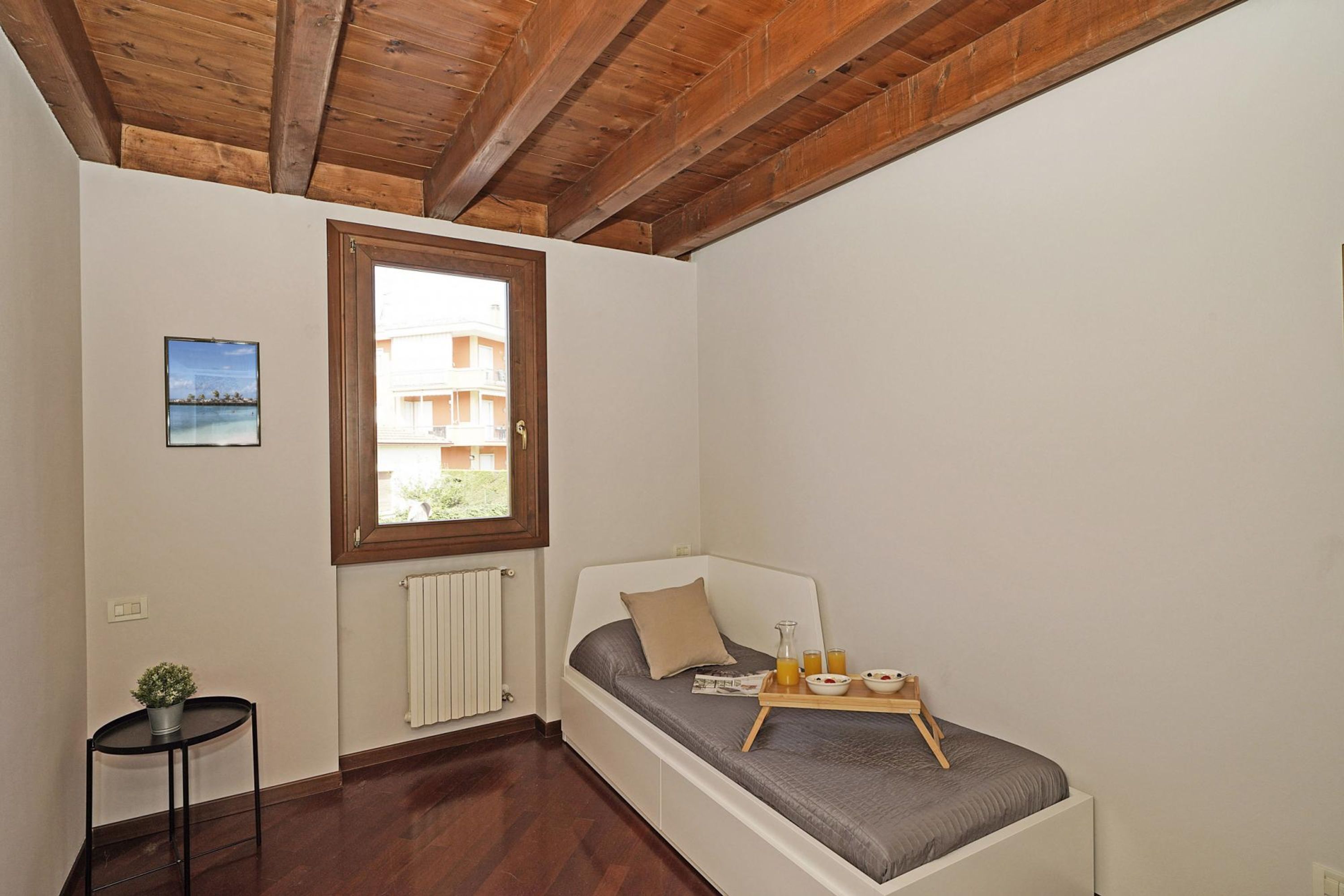 A small bedroom with a single bed, a window, a radiator, a small table with a potted plant, and a picture on the wall.