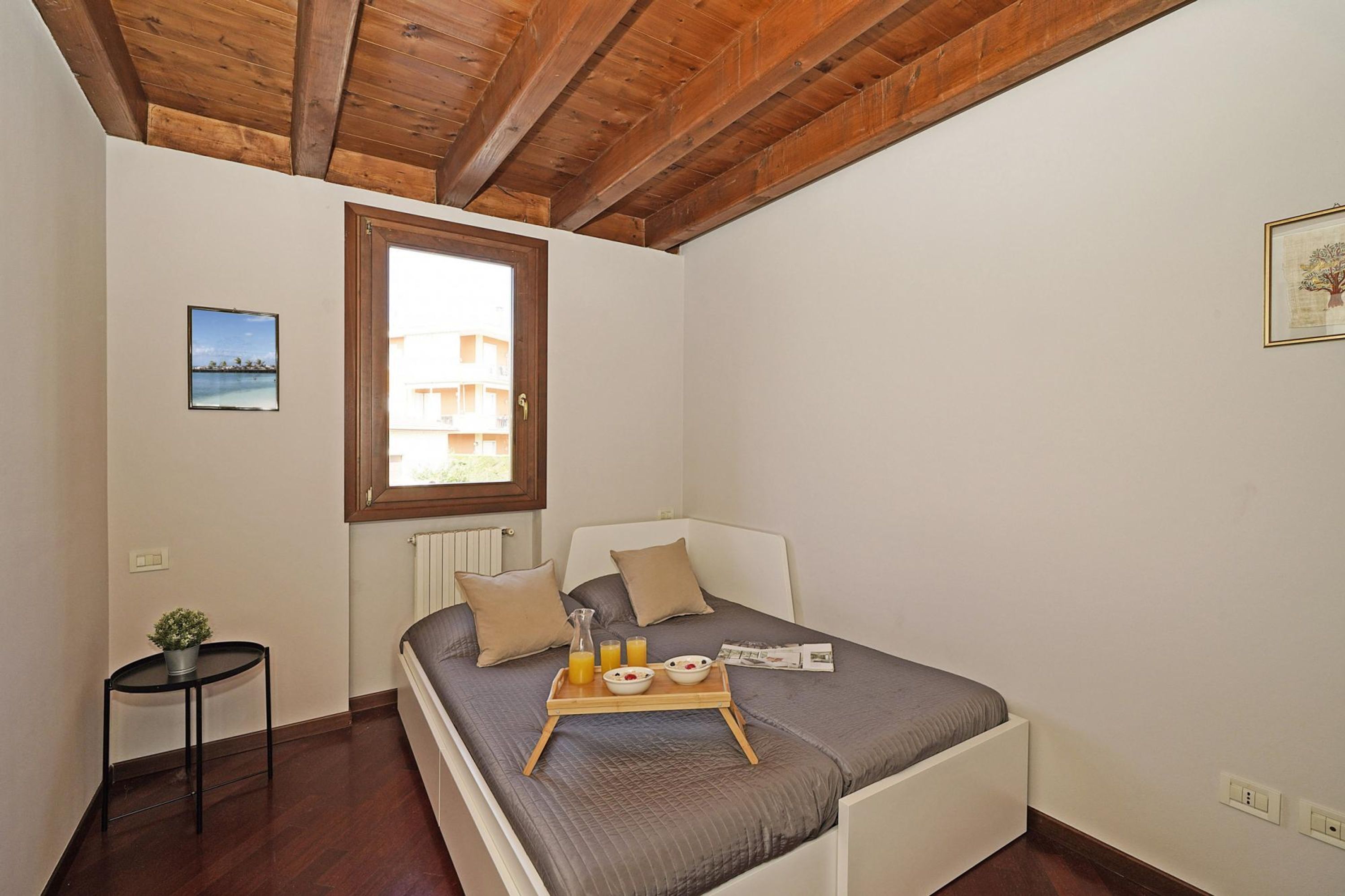 Un dormitorio con techo de madera, una cama con colcha gris, una mesita de noche con una planta y una ventana con vistas a edificios.