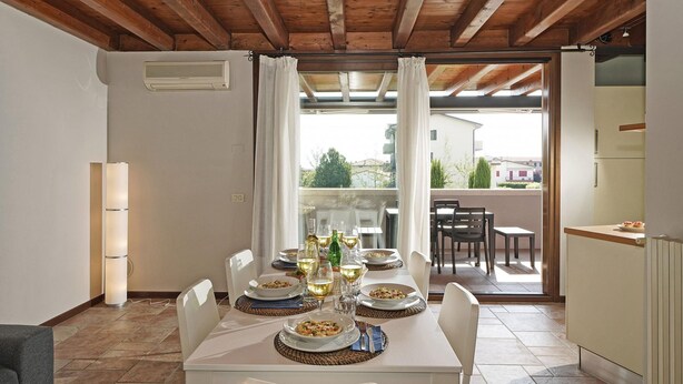 Uma sala de jantar com uma mesa posta para quatro pessoas, uma cozinha e vista para o exterior através de uma porta de vidro.