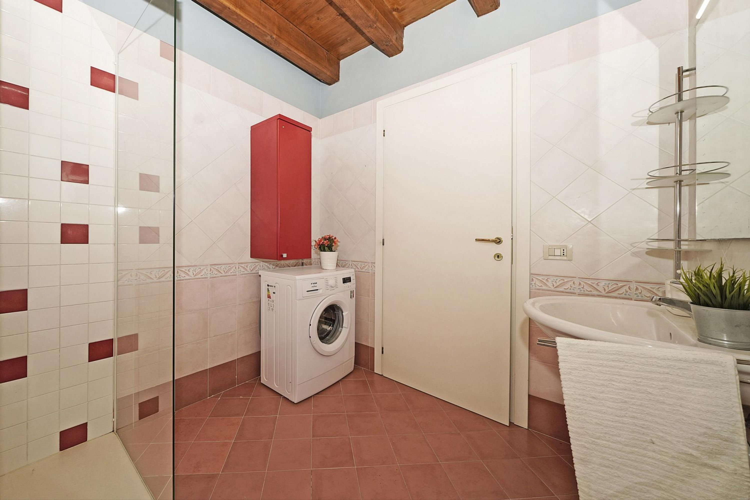 Ein kleines Badezimmer mit einer Waschmaschine, einem roten Schrank, einem weißen Waschbecken und einem Glastischchen mit einer Topfpflanze.