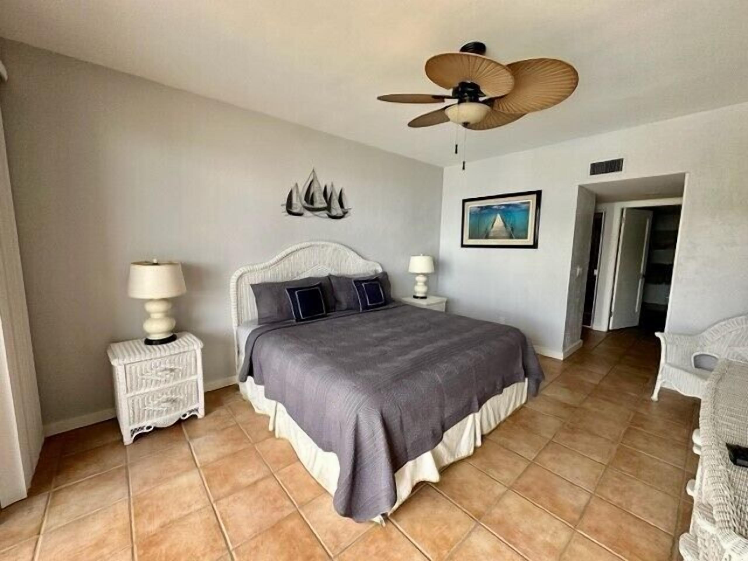 A bedroom with a large bed, two bedside tables with lamps, a ceiling fan, a wall-mounted sculpture, and a framed picture.