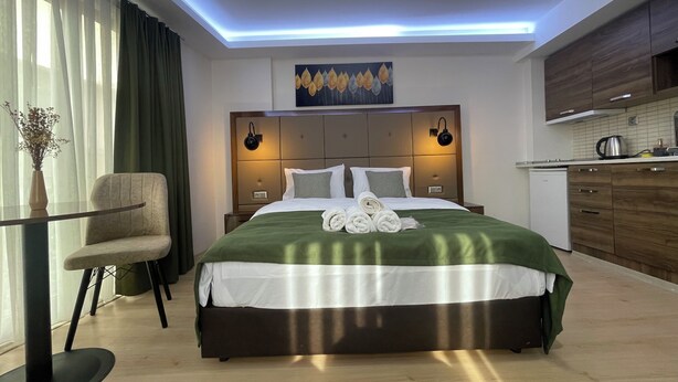 Una habitación de hotel moderna con una cama grande, lámparas de noche, una mesita con una silla, vista a la cocina y un cuadro en la pared.