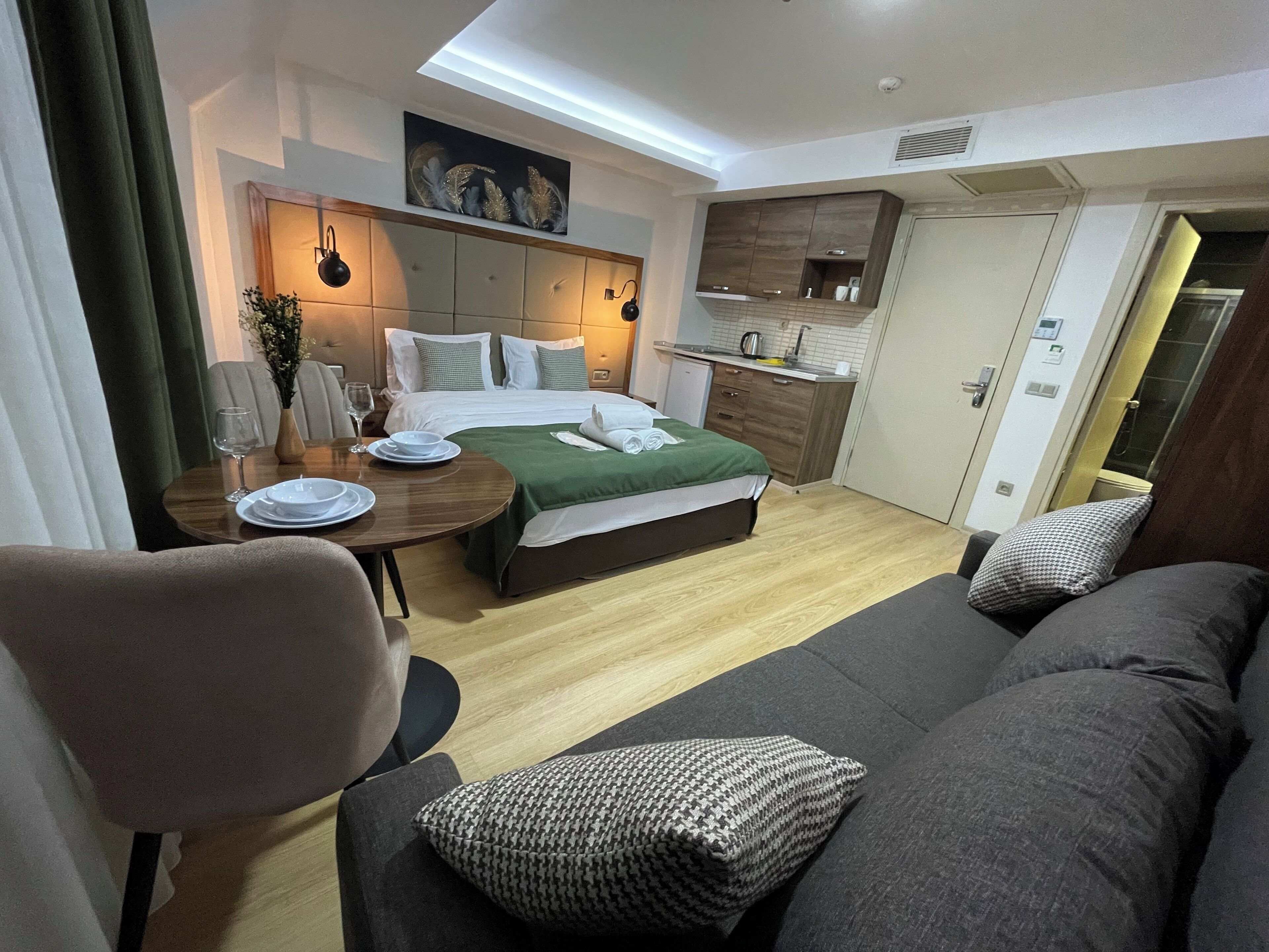 A modern hotel room with a bed, a dining area, a kitchenette, and a sofa.