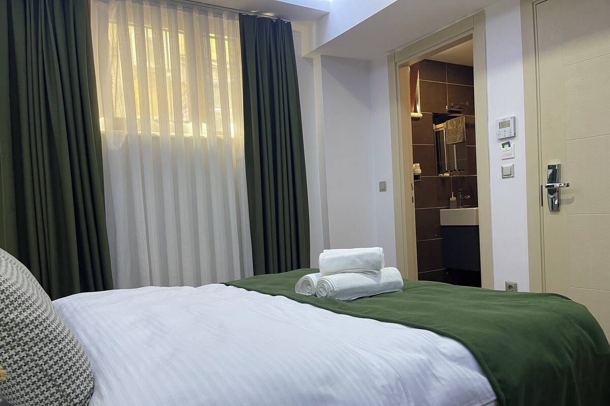 A hotel room with a bed, green and white curtains, a bathroom with a shower, and towels on the bed.