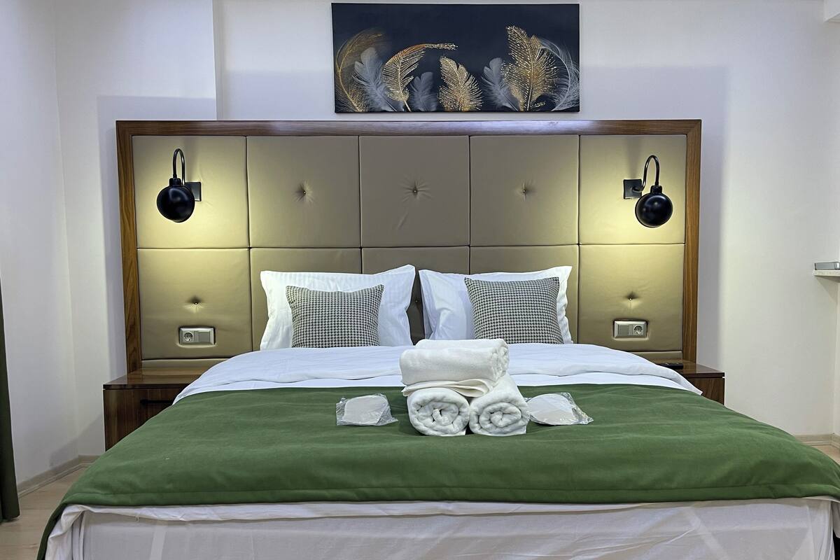 A neatly made bed with a green bedspread, white linens, and rolled towels. There are two bedside tables with lamps, a headboard with a decorative panel, and a framed artwork above the bed.