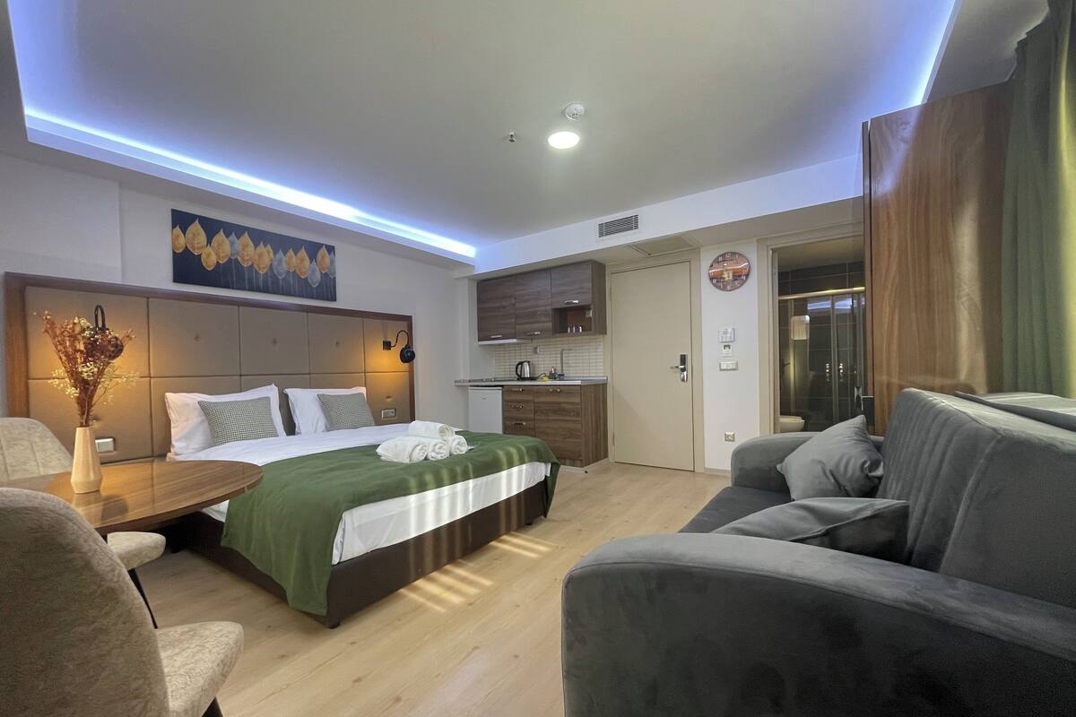 A modern hotel room with a bed, a sofa, a small table, a nightstand, a wall-mounted clock, and a kitchen area.