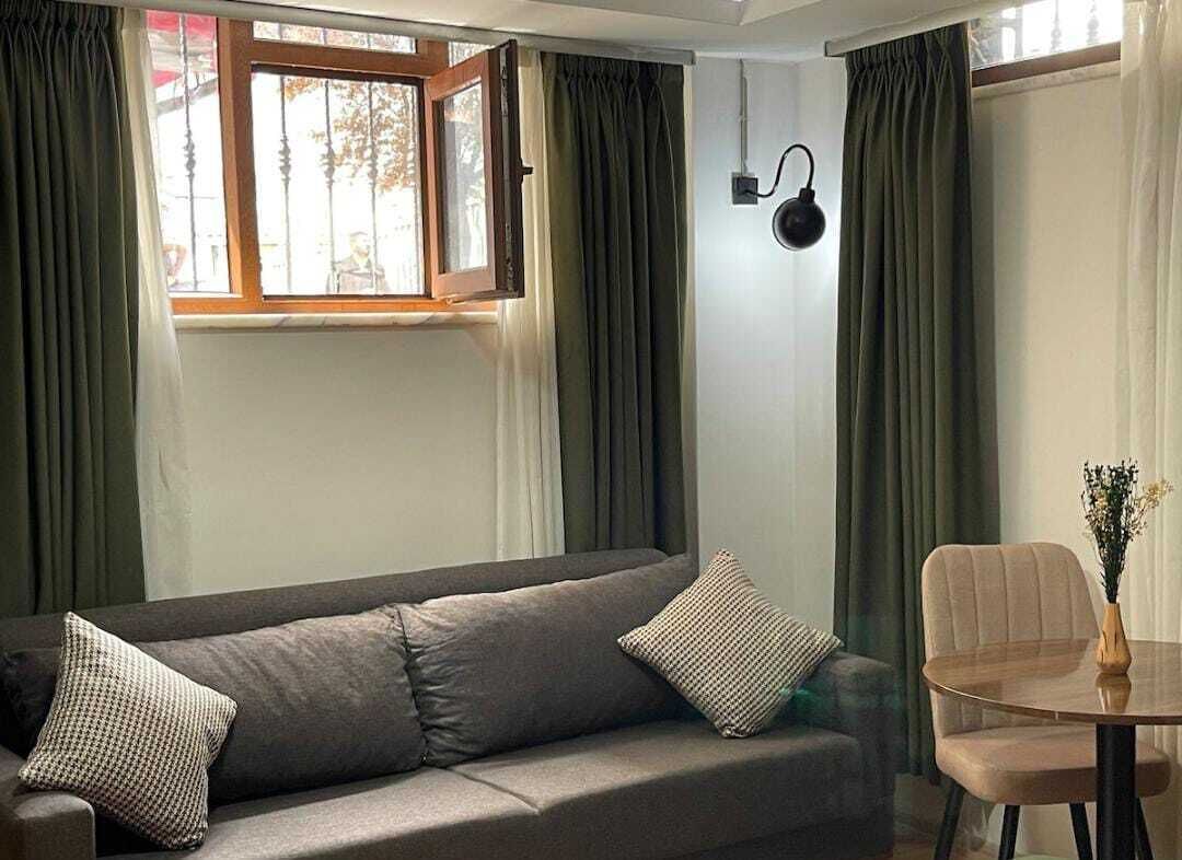 Een kamer met een bank, een eettafel met stoel, een raam met gordijnen en een wandlamp.