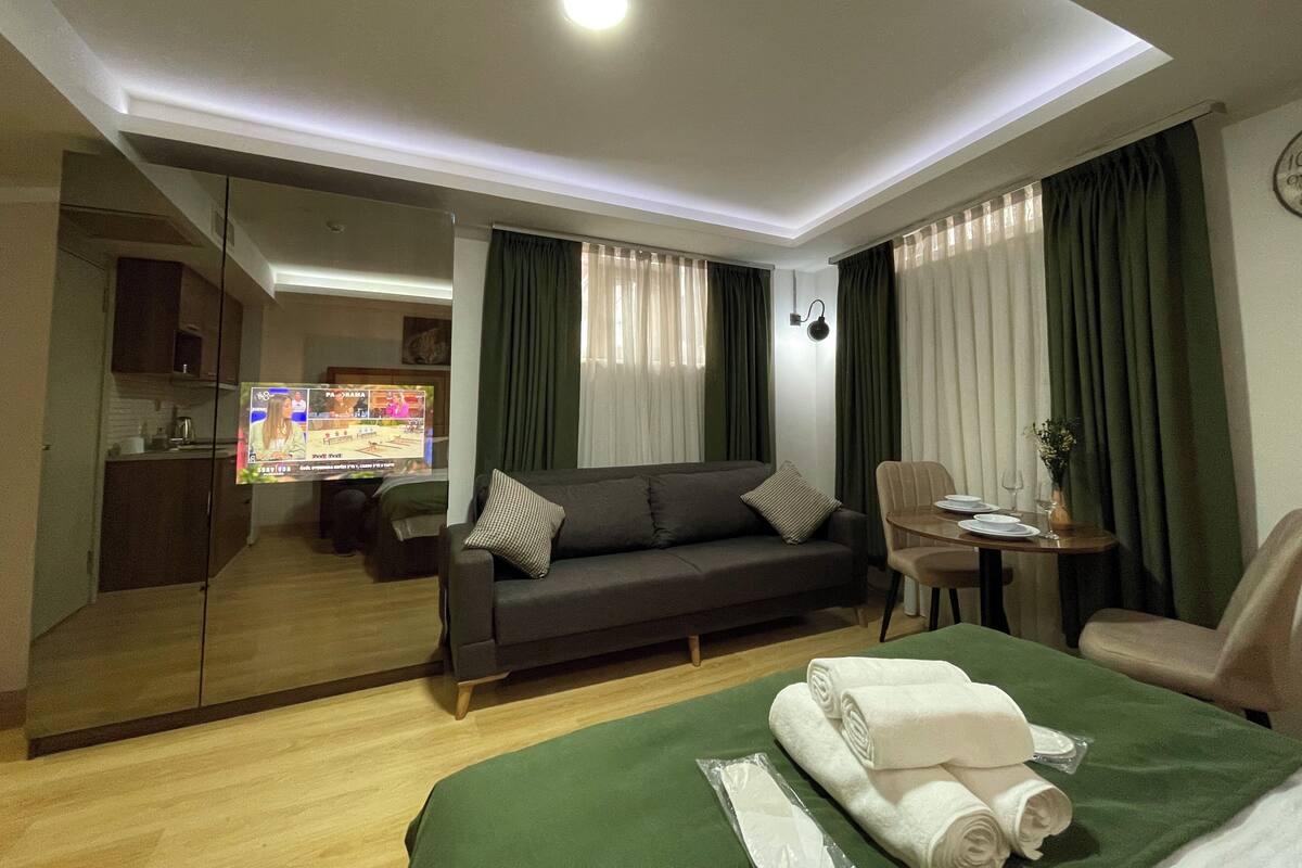 A hotel room with a sofa, a dining table with chairs, a small kitchen area, and a large mirror.