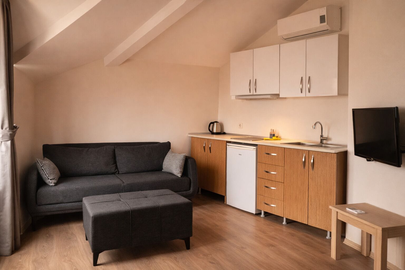 A compact living space with a sofa, ottoman, kitchenette, and a TV mounted on the wall.