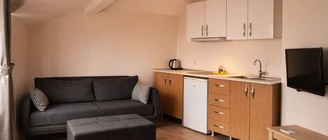A compact living space with a sofa, ottoman, kitchenette, and a TV mounted on the wall.