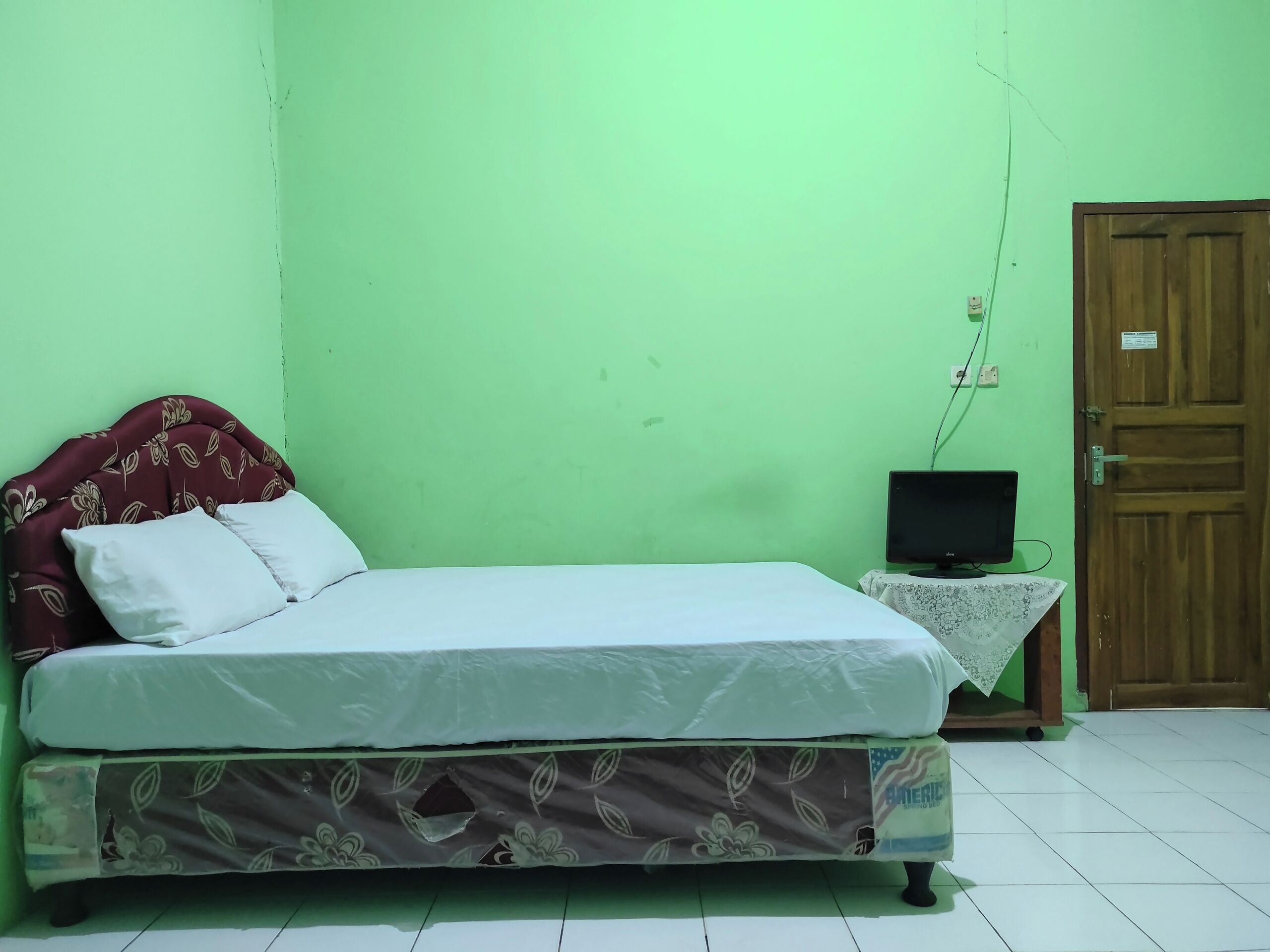 A single bed with a floral-patterned headboard and footboard, a small bedside table with a television, and a wooden door.