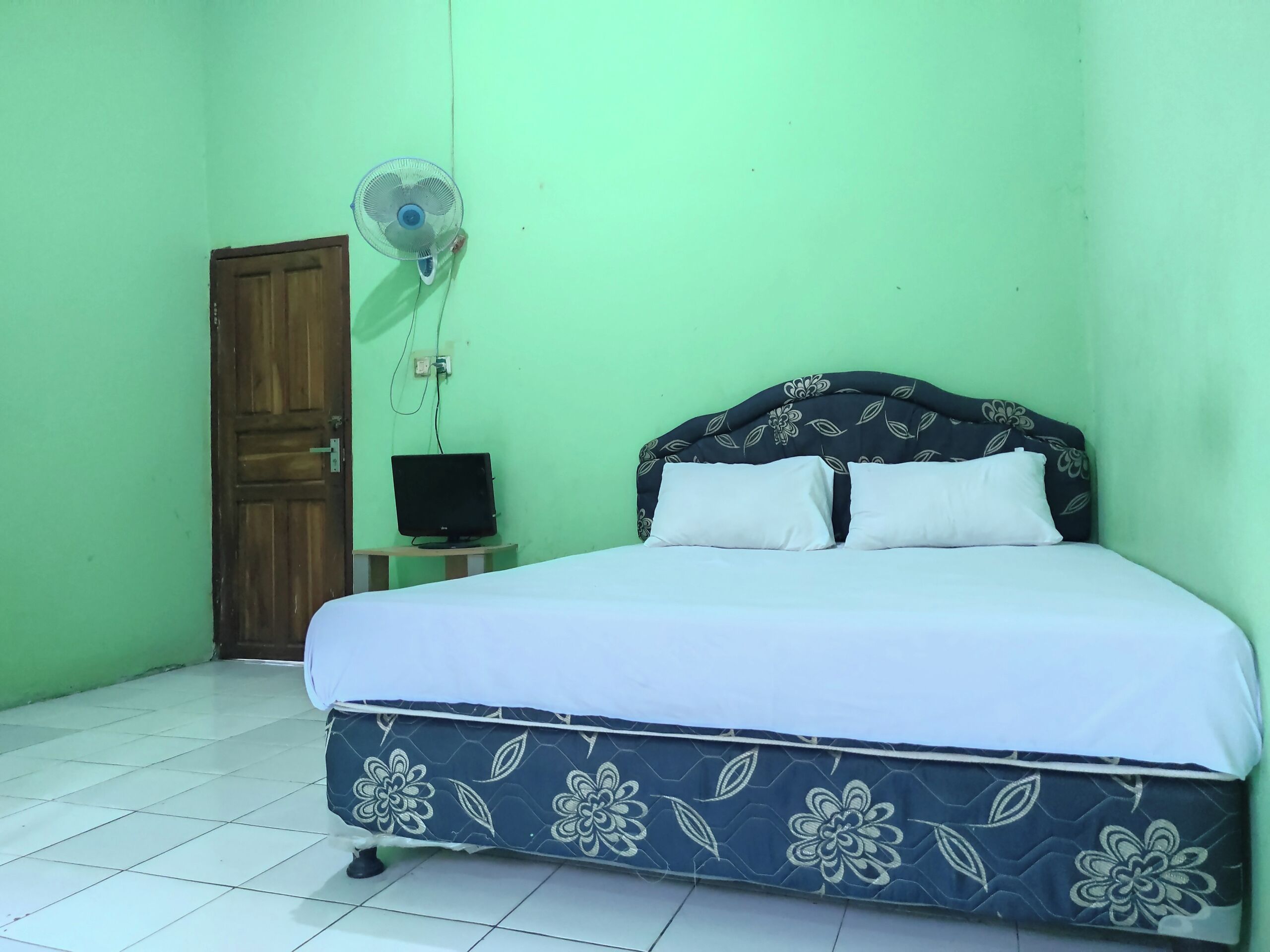 Un dormitorio con cama, un ventilador de pared, un televisor sobre una mesita y una puerta.