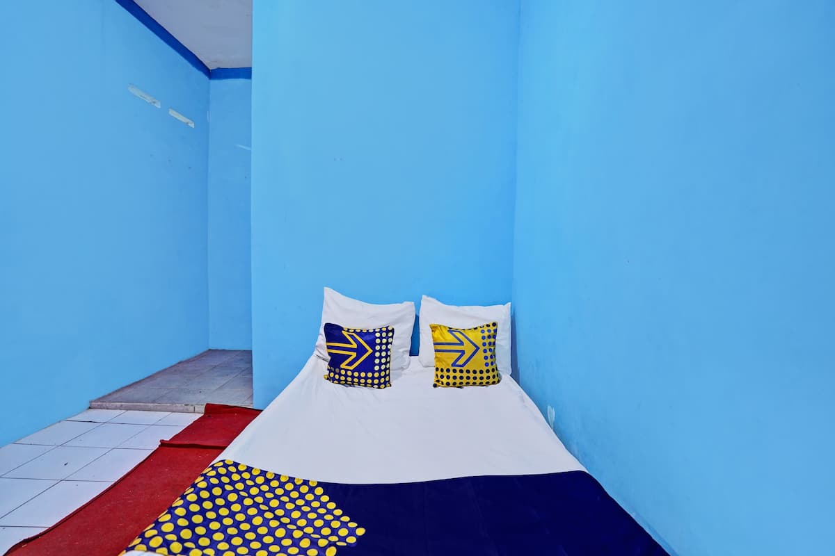 A small, brightly colored room with a single bed, two pillows with geometric patterns, and a red and yellow polka dot blanket.
