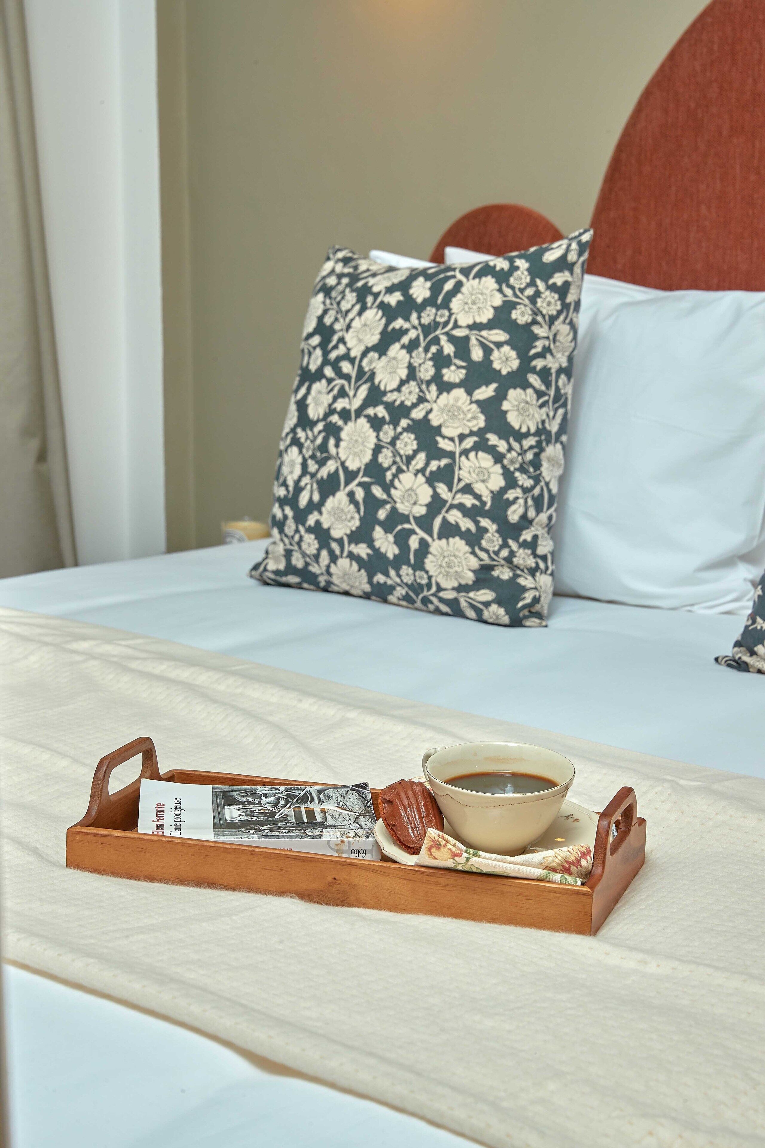 Una cama bien hecha con almohadas florales, una bandeja con una taza de café, un libro y una barra de chocolate.