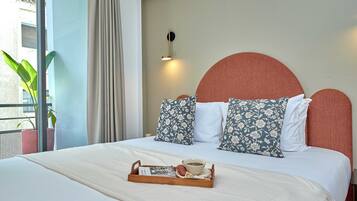 Una cama bien tendida con almohadillas estampadas, una bandeja con una taza y un libro, y vistas a un balcón con plantas.