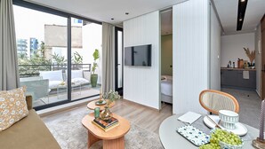 A modern living room with a sofa, coffee table, and dining area. There is a TV mounted on the wall and a balcony with outdoor furniture.