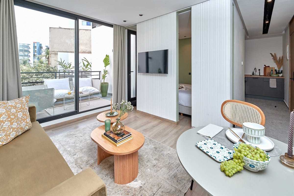 A modern living room with a sofa, coffee table, and dining area. There is a TV mounted on the wall and a balcony with outdoor furniture.