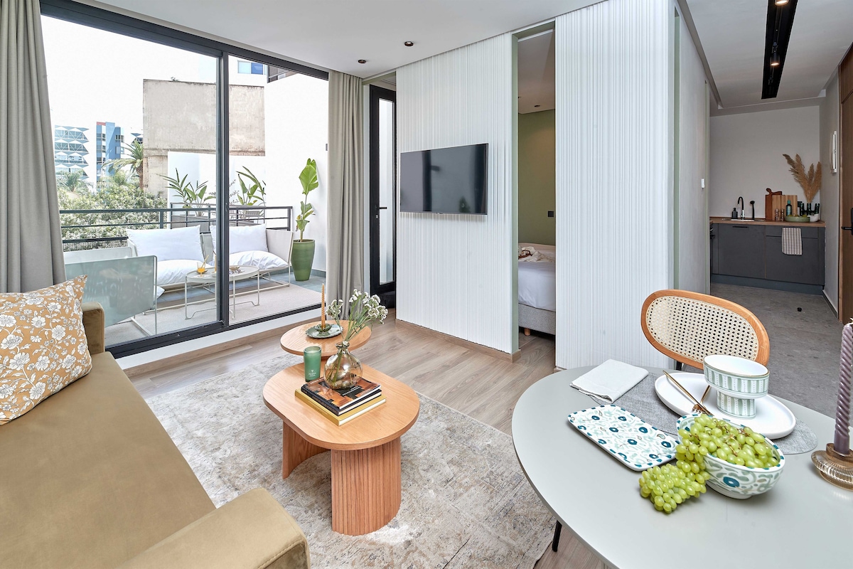 A modern living room with a sofa, coffee table, and dining area. There is a TV mounted on the wall and a balcony with outdoor furniture.