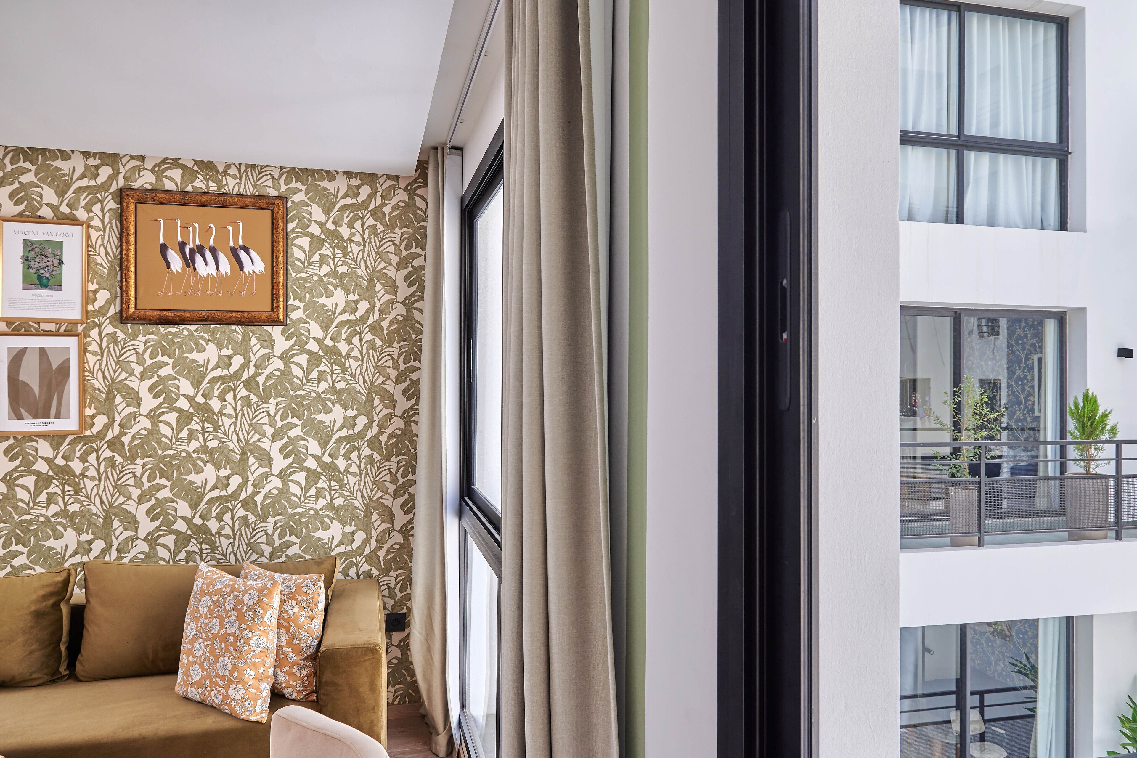 A room with a patterned wallpaper, a brown sofa with floral cushions, a framed artwork of birds, and a view of a balcony with plants.