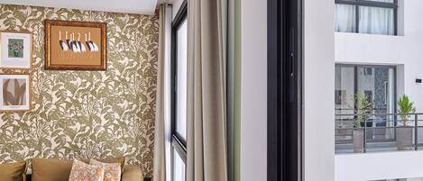 A room with a patterned wallpaper, a brown sofa with floral cushions, a framed artwork of birds, and a view of a balcony with plants.