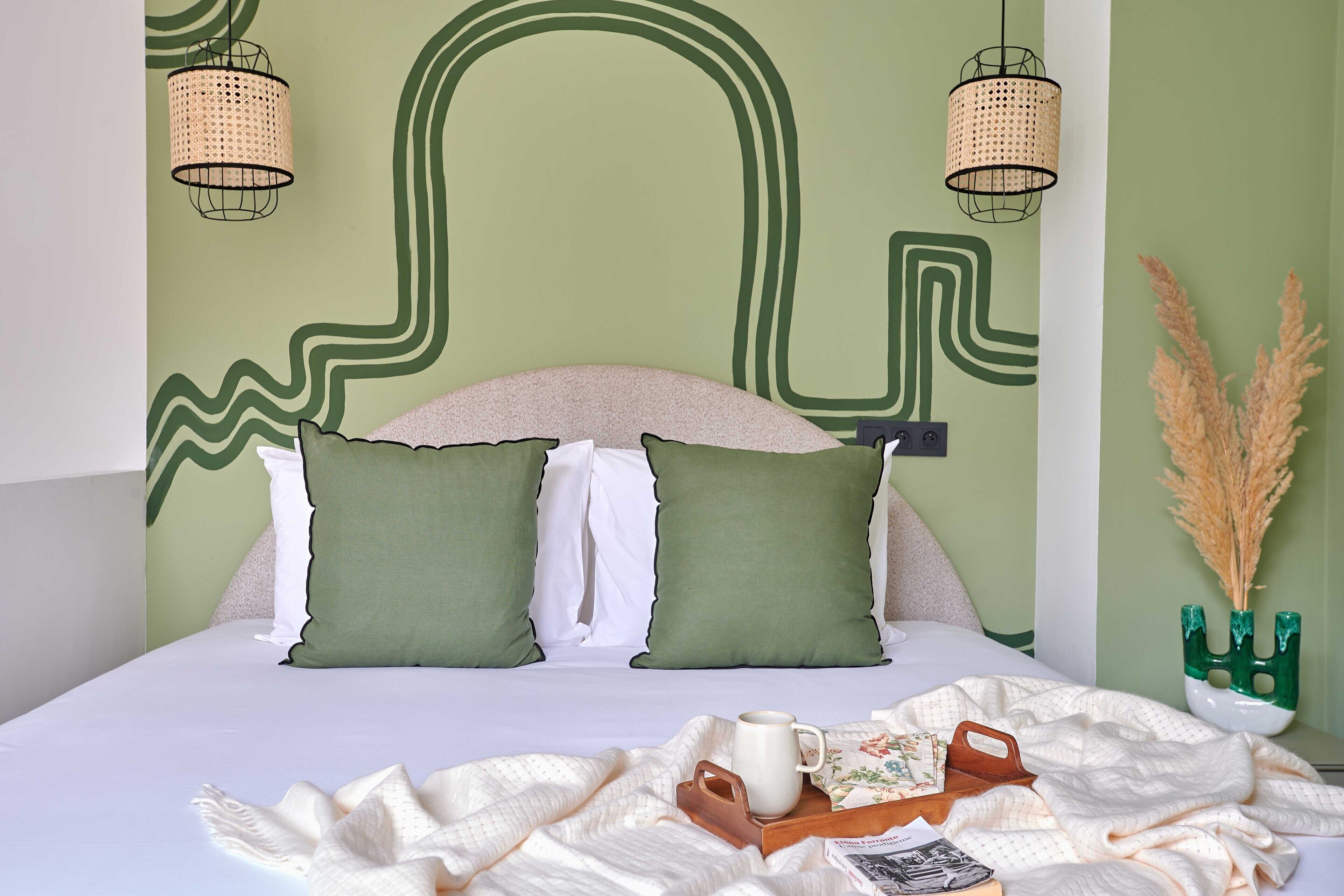 A bed with two pillows, a bedside table with a vase, and a wall with a green and white abstract design.