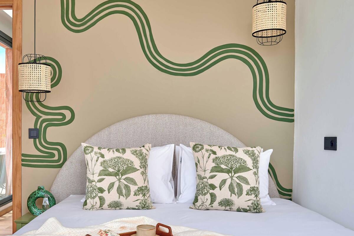 A bedroom with a curved headboard, two patterned pillows, a bedside table with a vase, and a window with curtains.