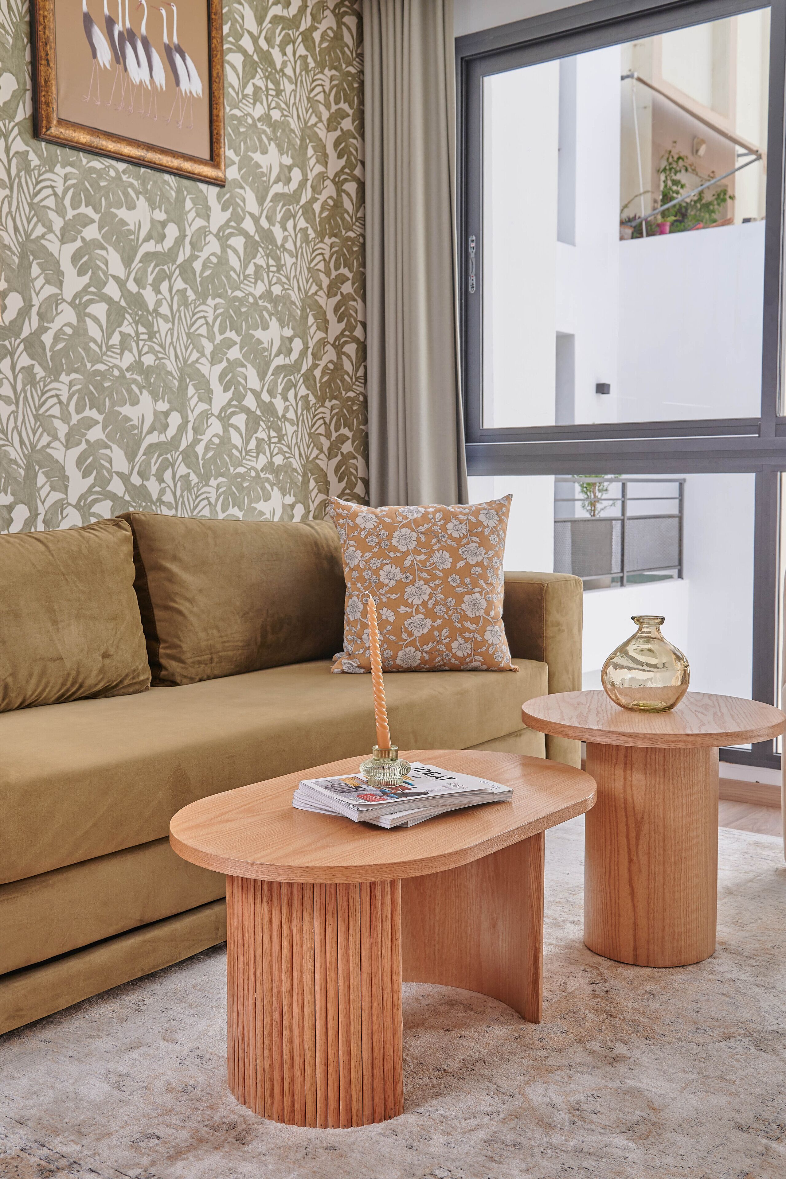 A living room with a brown sofa, a patterned pillow, two wooden side tables, and a large window with a view of a balcony.