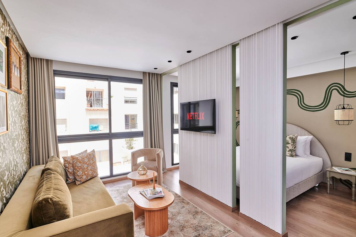 A modern hotel room with a sofa, a small table, a TV, a bed, and a view of the outside.