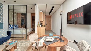 A modern hotel room with a dining area, a bed with blue and white bedding, a TV mounted on the wall, and a sliding door leading to a bathroom.