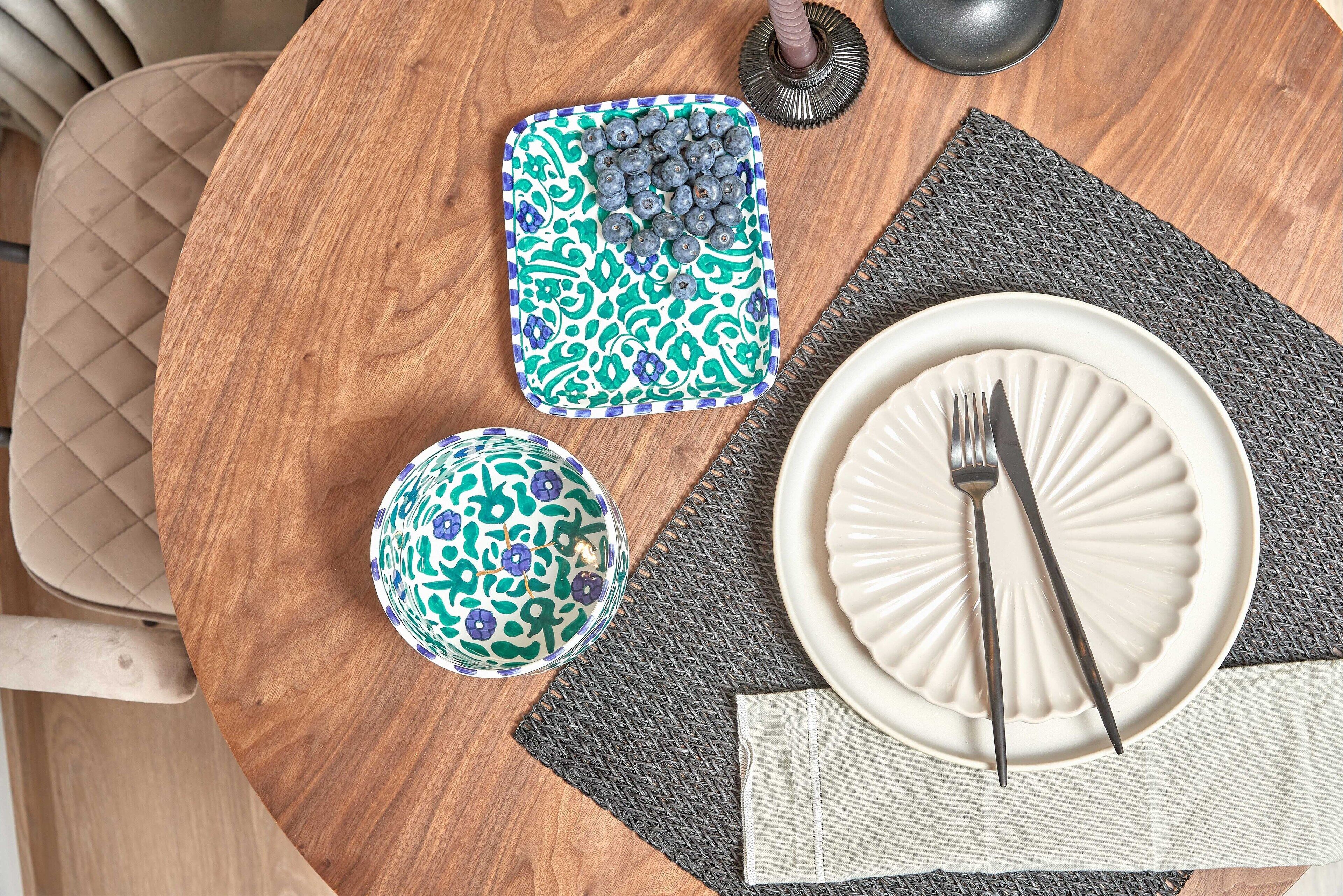 A round wooden table with a white plate, a fork, a bowl, and a patterned tray.