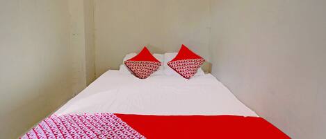 Ein kleines Zimmer mit einem Einzelbett, einer schlichten weißen Wand und einer rot-weißen Bettdecke.